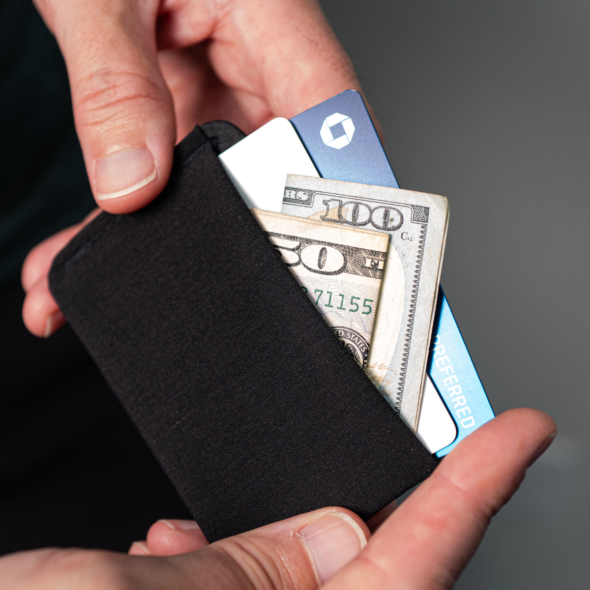 A close-up of a black fabric minimalist card holder wallet being held in two hands, with a U.S. all-groups