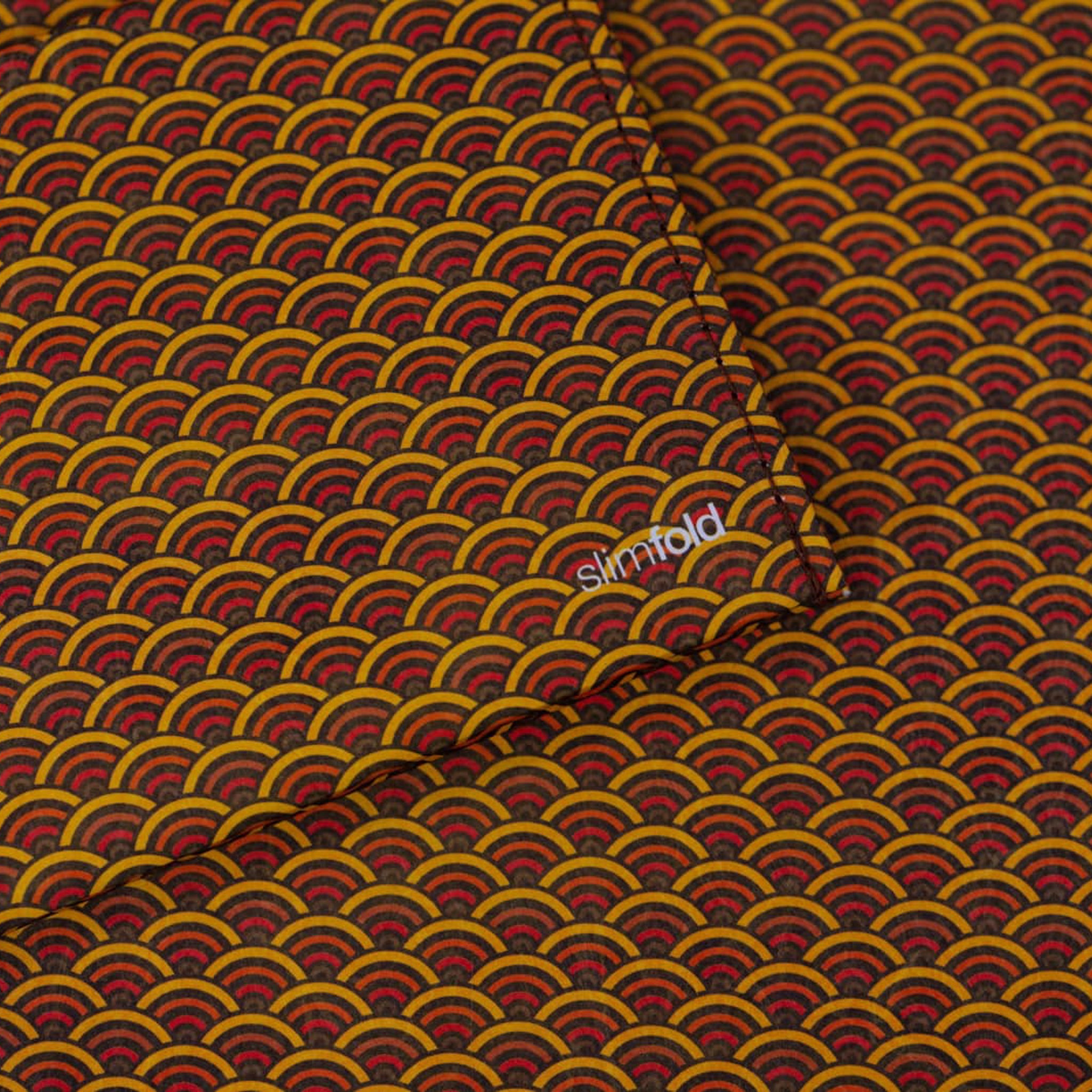 A close-up of a SlimFold Micro Tyvek wallet with a repeating Japanese-style wave pattern in mustard yellow, red, and dark brown, with a small white 'simfold' logo printed in the center.