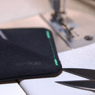 A close-up of a black fabric wallet with a green stitched edge and a debossed 'surfold' logo, resting on a white surface near a sewing machine and scissors. all-groups