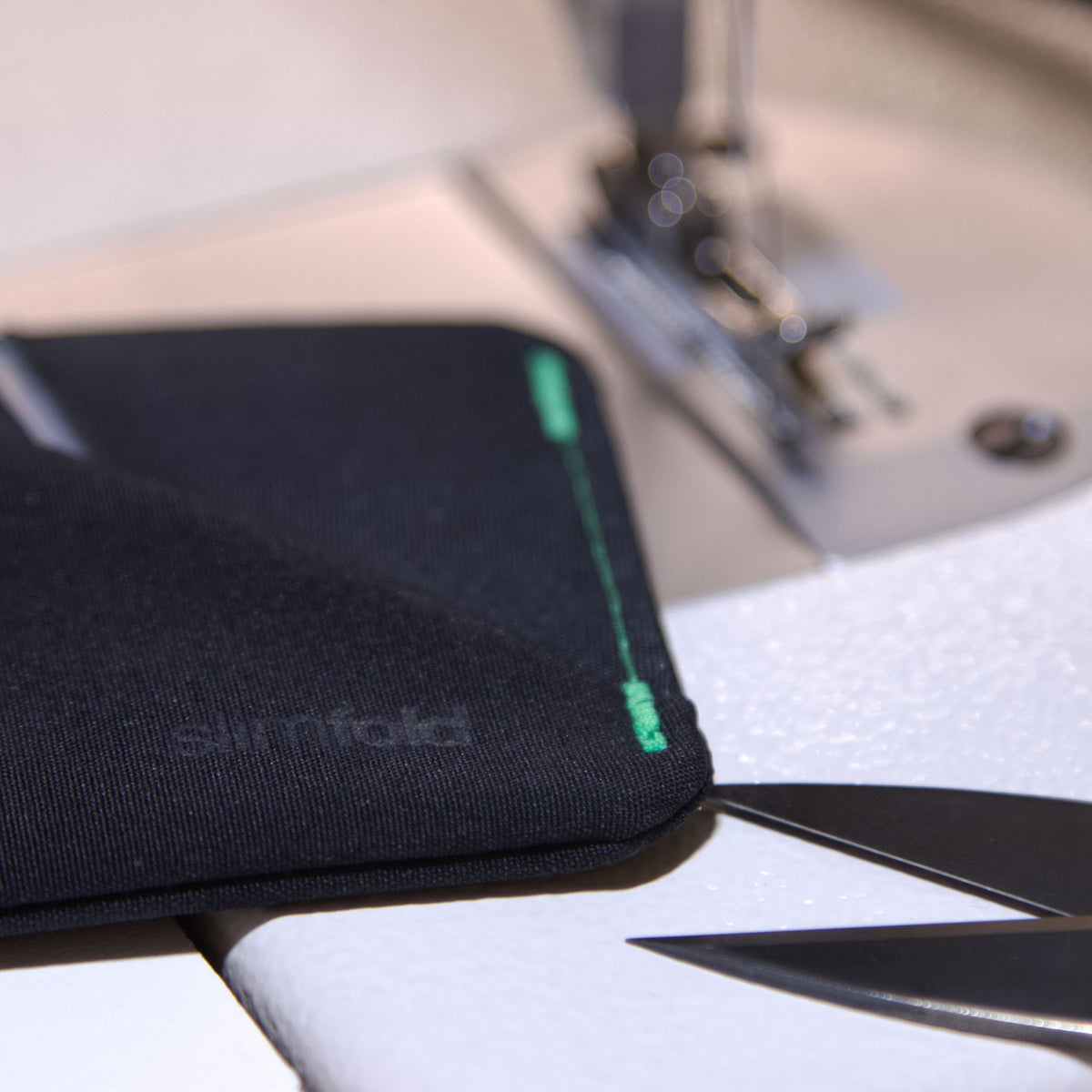 A close-up of a black fabric wallet with a green stitched edge and a debossed 'surfold' logo, resting on a white surface near a sewing machine and scissors. all-groups