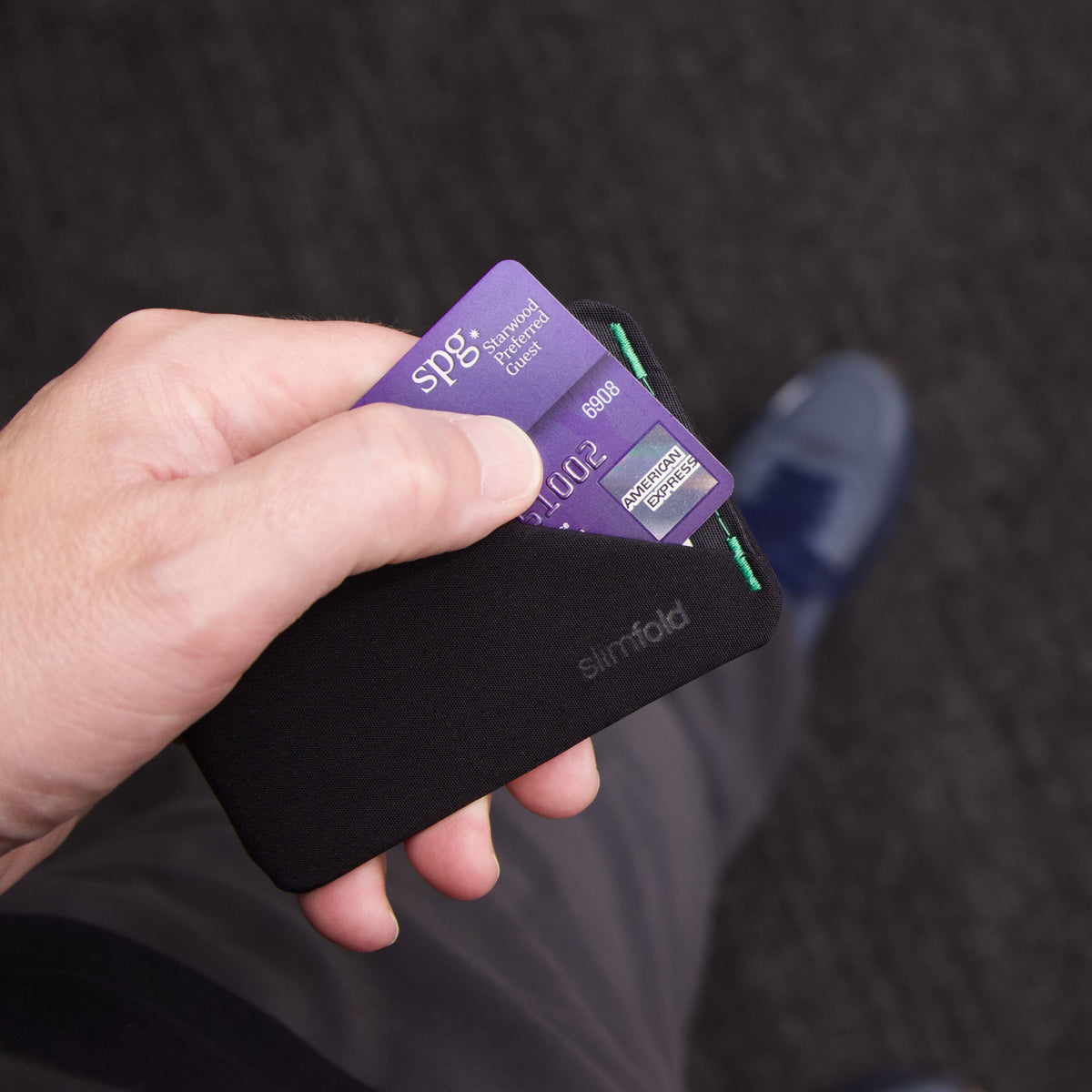 A close-up of a hand holding a black Slimfold card holder with two cards partially inserted, against a dark carpeted floor. all-groups