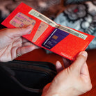 A close-up of a person holding an open red wallet with multiple card slots containing credit cards and cash. The wallet has a textured red pattern and is shown in use against a blurred background.