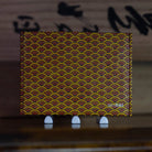 A thin SlimFold Micro Tyvek wallet with a repeating Japanese-style wave pattern in yellow, red, and dark brown, displayed on a wooden surface with three small white stands. The brand name 'simfold' is printed in the bottom right corner.